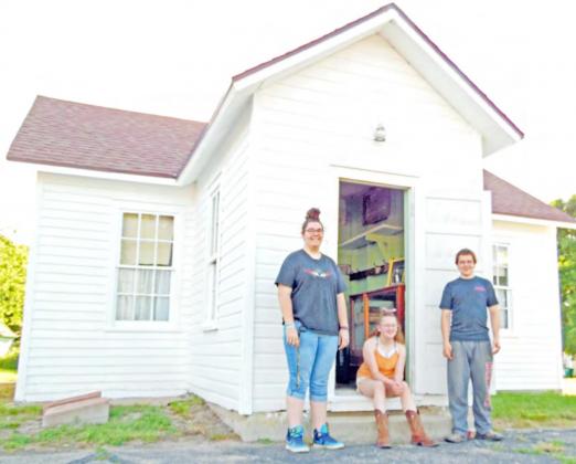 Historical Society volunteers Larissa Weimers, Colby Hedden, &amp; Zack Hedden stand at the door of the Maple Wood Country School Museum in Table Rock. The school originally sat near the intersection of what became Highways 50 &amp; 65. It was built in 1874, almost 20 years after Lydia Griffing taught 11 students in her 11 x 13’ log cabin.